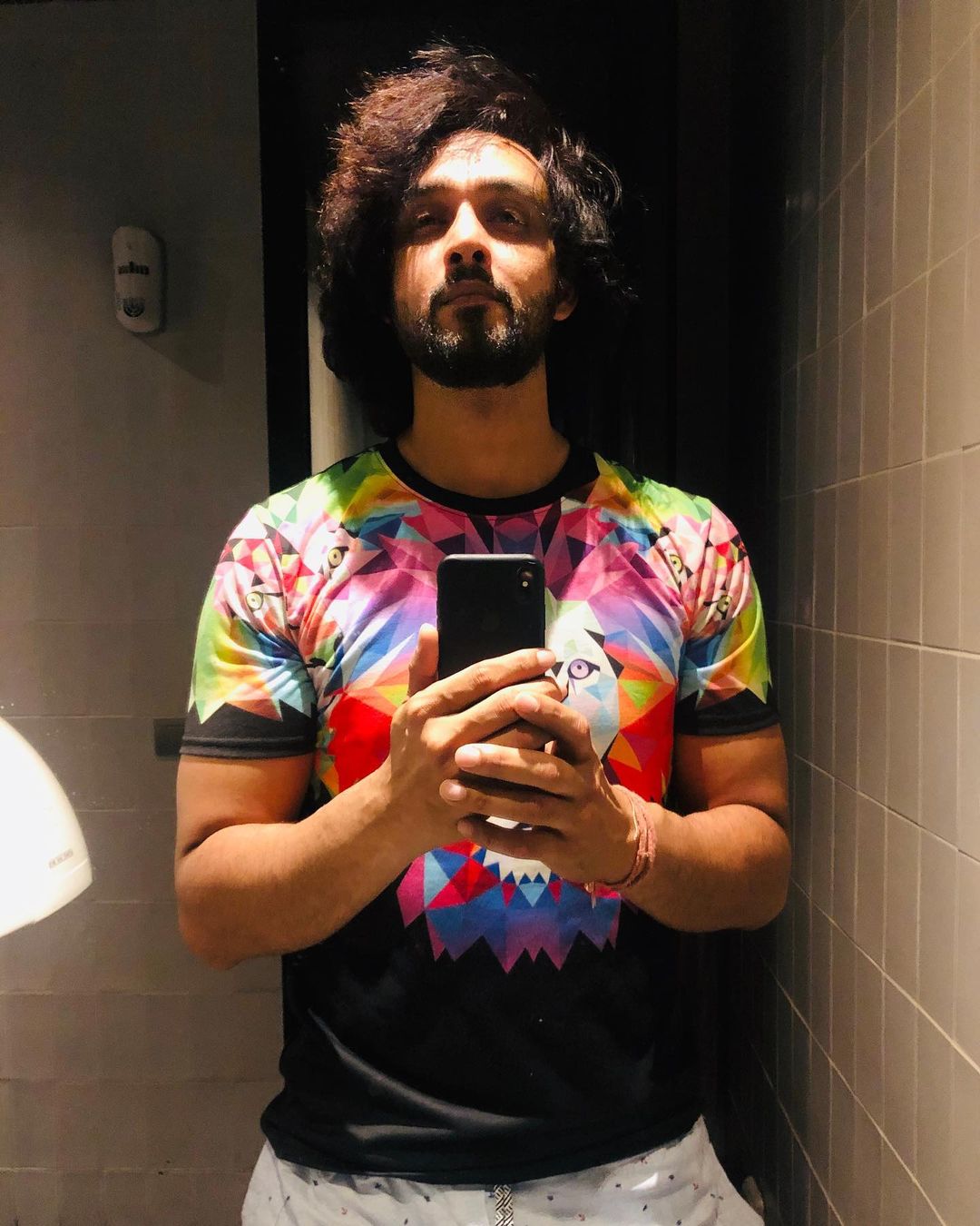 Sourabh Raaj Jain snaps a mirror selfie in October 2019. The room is dark, with mushroom coloured tiles, and is lit only from directly above and to one side. Sourabh has long dark curly hair and a thick, trimmed beard and moustache, and is wearing a black and multicoloured t-shirt and white patterned trousers. He is looking into the mirror.