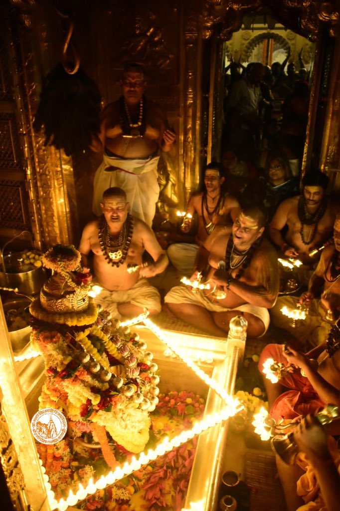 In a small, dark room, seated priests from each of seven lineages perform a ritual fire offering to a lingam, an abstract form of the Hindu god Shiva said to represent his manifestation as an infinite pillar of light. The lingam is decorated with garlands of flowers and gold accessories, and surrounded by lit ghee lamps.
