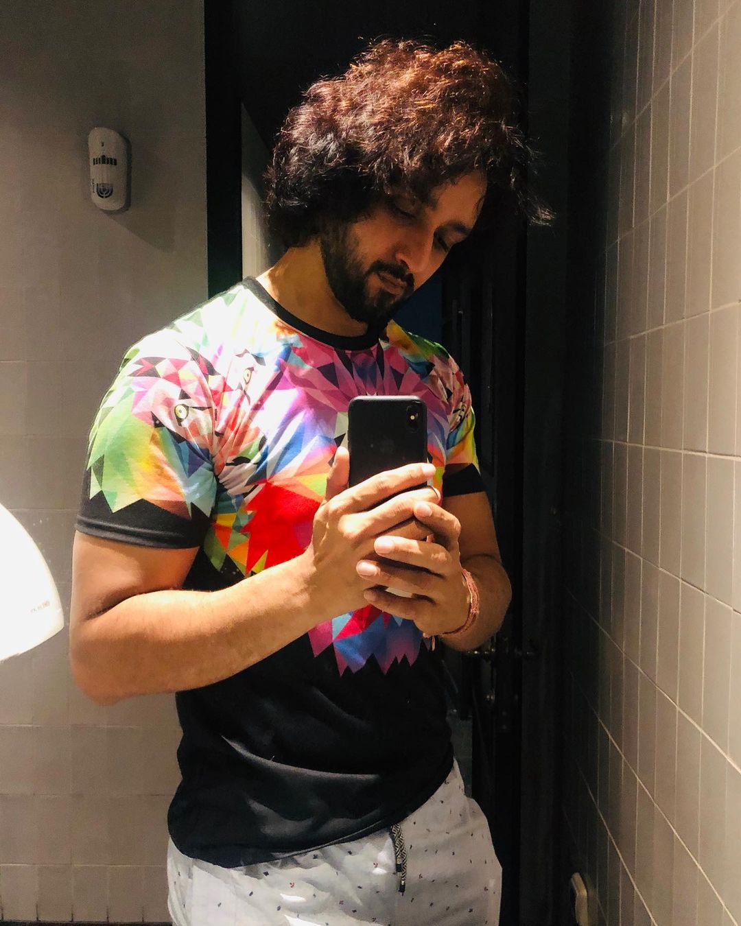 Sourabh Raaj Jain snaps a mirror selfie in October 2019. The room is dark, with mushroom coloured tiles, and is lit only from directly above and to one side. Sourabh has long dark curly hair and a thick, trimmed beard and moustache, and is wearing a black and multicoloured t-shirt and white patterned trousers. He is looking at his smartphone.