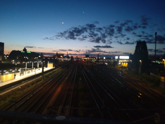 Foto einer Morgendämmerung in der Stadt (Berlin). In der Mitte liegen Gleise im Dunkeln, die vom unteren Bildrand bis zum Horizont in der Bildmitte führen. Links ein verwaschener S-Bahn-Bahnhof. Rechts, links und im Hintergrund Gebäude. Am Morgenhimmel klein die Mondsichel und ein Stern erkennbar. Vereinzelte Wölkchen. 