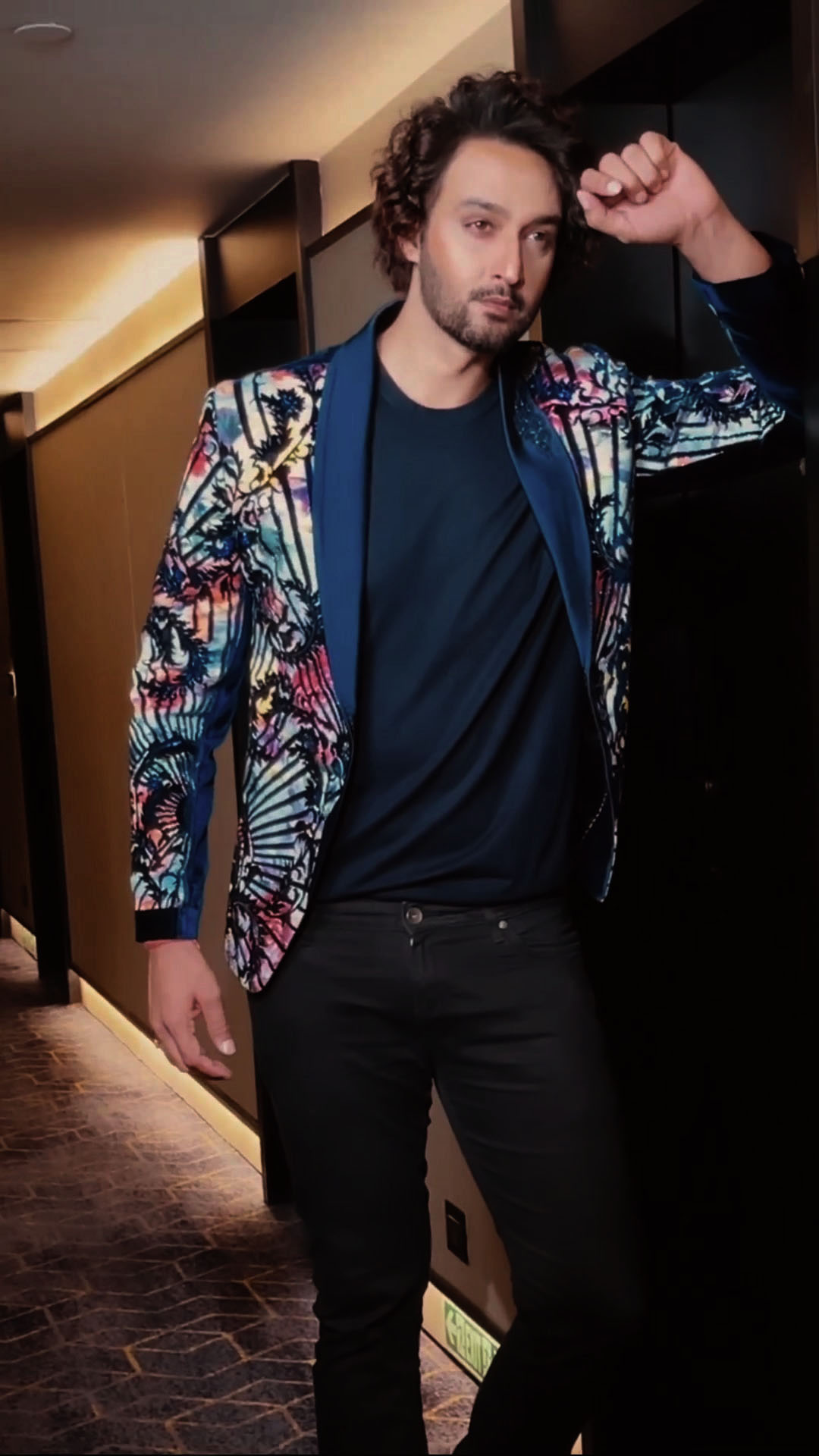 Sourabh Raaj Jain is seen posing during a photo shoot in Mumbai in autumn 2022. He has dark brown, medium-length hair and a trimmed beard and moustache. He is stood in a low-lit hallway and is wearing a flamboyant, high-contrast multicoloured tuxedo in a floral baroque design, a navy t-shirt and black jeans. He is leaning on a doorway while looking directly at the photographer's camera, and is captured in this image using a second camera.