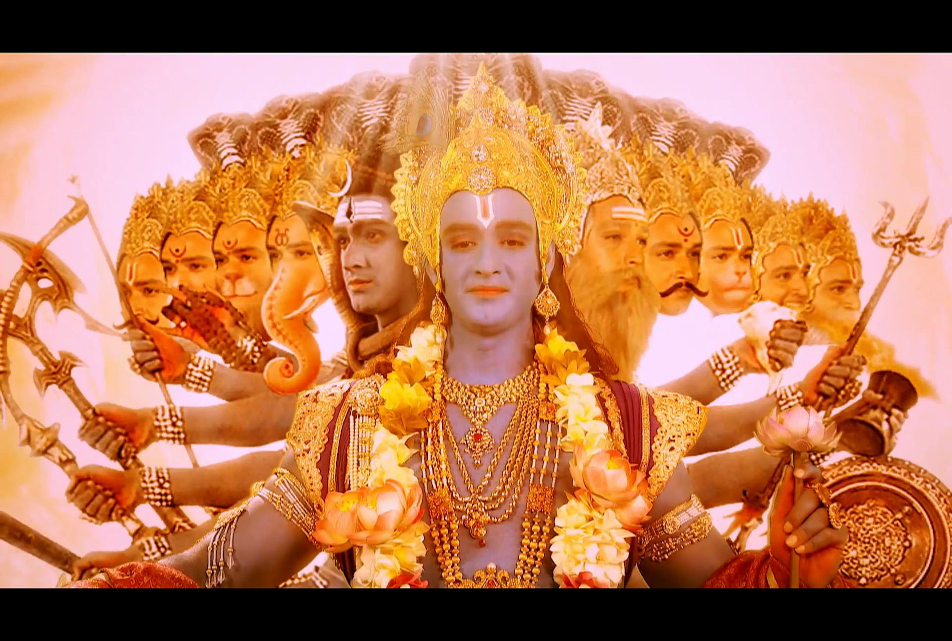 Sourabh Raaj Jain plays Shri Krishna in the Vishwaroop scene of the Bhagavad Gita in Star Plus Mahabharat (2014), in which he reveals to the warrior prince Arjun the knowledge of his universal form. Krishna is seen as the supreme form of the Hindu god Vishnu, decked in fine, gold-embellished garments, gold jewellery and an elaborate gold mukut (tall crown), draped in a garland of flowers and bathed in golden light. The faces and arms of his many avatars extend either side of him, weapons in hand, and the many-headed snake demigod Shesha is visible behind him. He looks down at Arjun with an expression of calm benevolence.