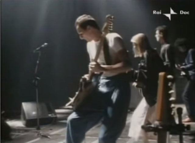 stage photo of a talking heads concert in rome in 1980. from left front to right back: adrian belew guitar, tina weymouth bass, david byrne vocals and guitar