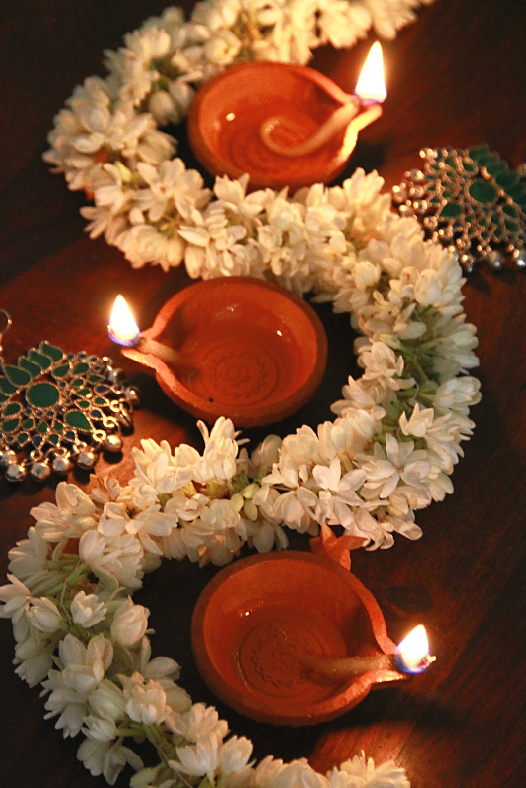 A garland of delicate white jasmine flowers and decorations of green enamel and gold arranged around three lit diyas (oil lamps) made of terracotta