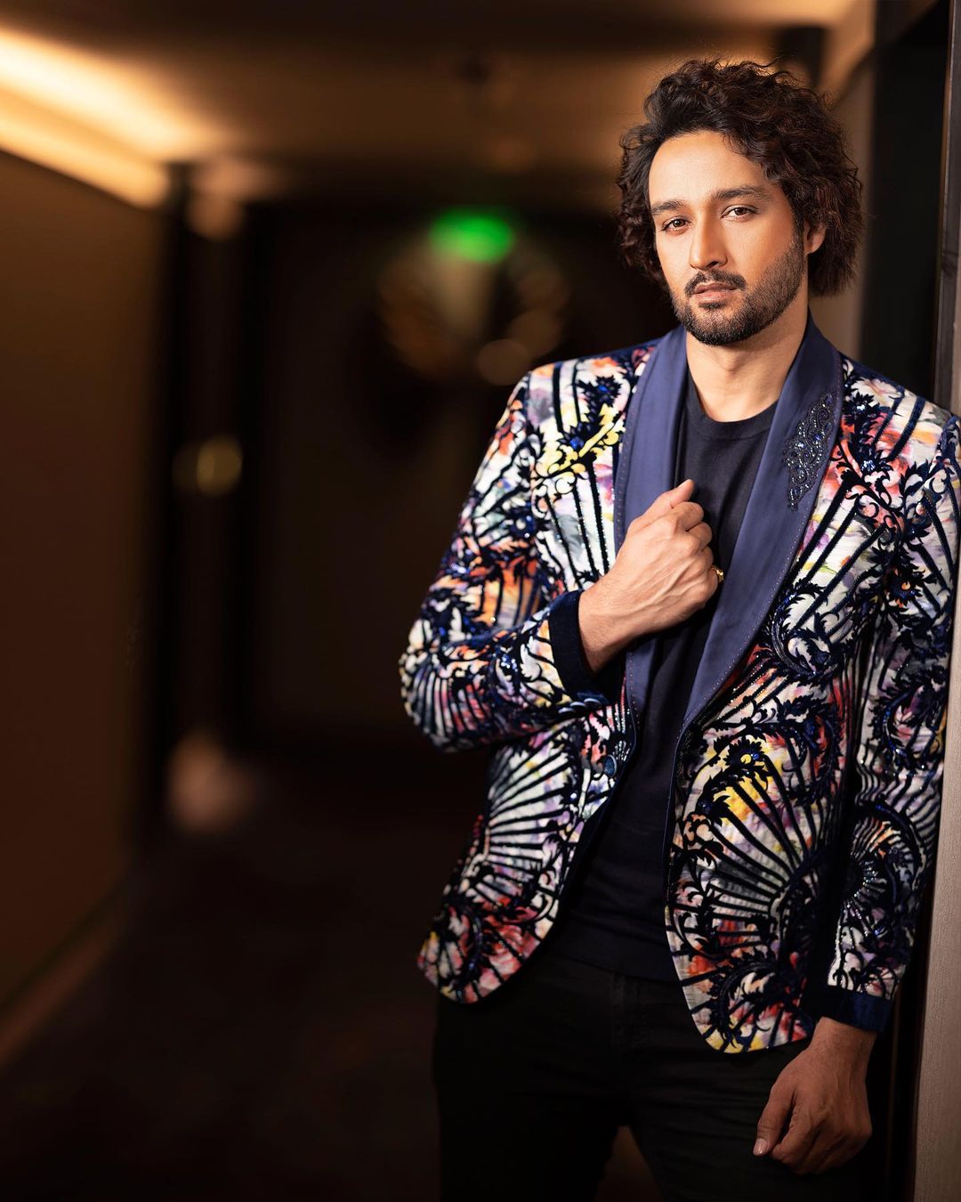 Sourabh Raaj Jain poses for a photo in Mumbai in autumn 2022. He has dark brown, medium-length curly hair swept to one side and a trimmed beard and moustache. He is stood in a low-lit hallway and is wearing a flamboyant, high-contrast tuxedo made from pale fabric with a vibrant, multicoloured floral motif that is embellished with black appliqué and blue beadwork in a baroque tendril design, with a dark blue satin collar that also features beadwork on one side. The jacket is styled with a plain navy blue t-shirt and black jeans. He is looking directly at the camera.