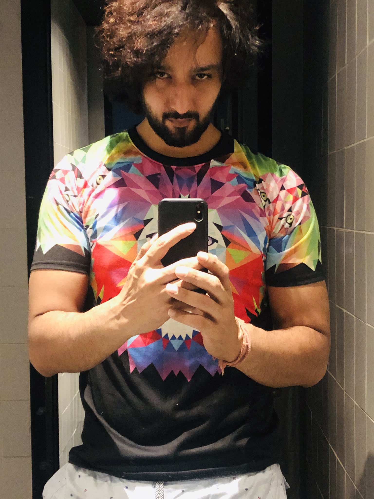 Sourabh Raaj Jain snaps a mirror selfie in October 2019. The room is dark, with mushroom coloured tiles, and is lit only from directly above and to one side. Sourabh has long dark curly hair and a thick, trimmed beard and moustache, and is wearing a black and multicoloured t-shirt and white patterned trousers. He is looking directly at the camera.