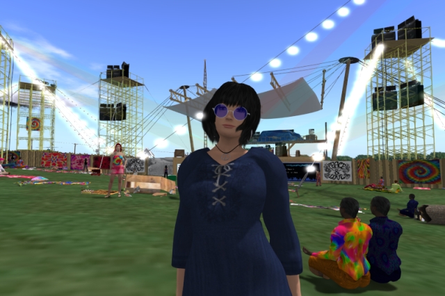 Self-portrait in a hippie look on the Woodstock festival grounds; I'm wearing tinted wire-framed glasses, a long blue dress and a peace necklace; behind me, there is the stage with its two PA towers, separated from the audience by a wooden fence with posters on it