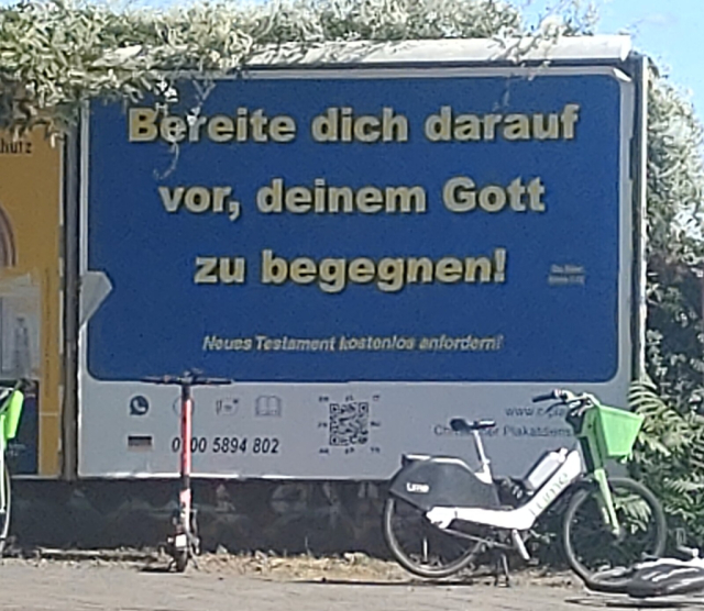 Ein Werbeplakat. Großes, rechteckiges blaues Feld auf weißem Grund. Im blauen Feld ein Text in gelber schrift: Bereite dich darauf vor, deinem Gott zu begegnen. Anrede klein geschrieben. Im weißen Bereich am unteren Rand links eine Deutschlandfahne, rechts davon eine Telefonnummer, rechts davon ein QR-Code. Davor steht ein Mietrad und ein Mietroller