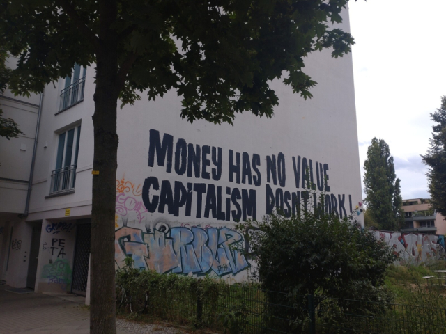 mural inscription, black capital letters on a white wall of a house behind a tree and some shrubbery: MONEY HAS NO VALUE CAPITALISM DOSNT WORK ! below some indecipherable mural tags.