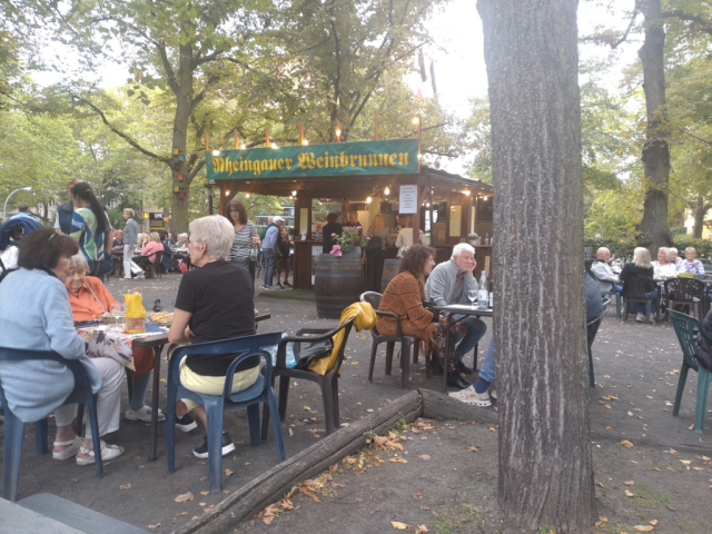 ausschank des rheingau weinbrunnens in berlin am rüdesheimer platz. unter bäumen mit leuten auf gartenstühlen an tischen