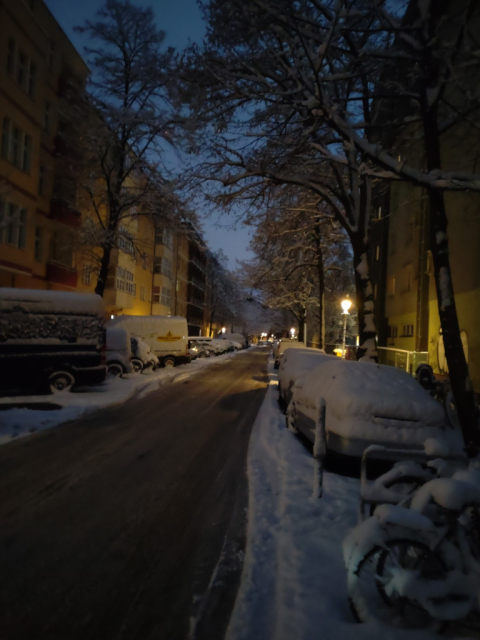 verschneite straße in berlin mit parkenden autos zu beiden seiten. morgendämmerung. zwischen den parkenden autos und den häusern stehen kahle bäume, die äste mit schnee bedeckt, die autos ebenfalls. der schnee auf der straße ist schon zu matsch geschmolzen.